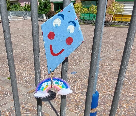 Foto di una sagoma di aquilone sorridente, alla quale è appeso un arcobaleno, attaccato al cancello della scuola SP Aldeno